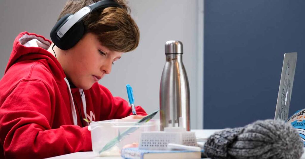 Boy sitting at a desk with headphones on studying inb front of a computer laptop