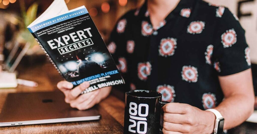 Man in a short sleeve shirt with a book in one hand and a coffee cup in the other with an 80 20 rule on it
