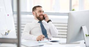 smiling man in a suit sitting a desk holding a mobile phone to his ear