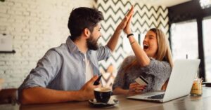 Couple at a table high fiving while sitting in front of a laptop in a coffee house