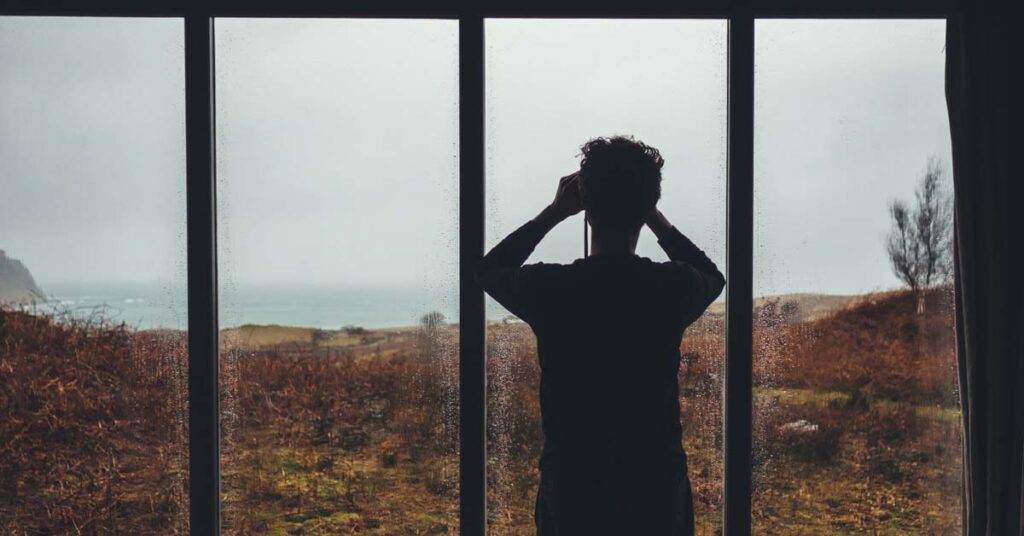 Man standing at a glass window looking through binoculars to the sea