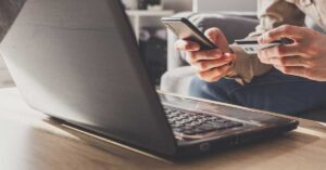 Laptop on a table with hands holding a mobile phone and credit card in front it