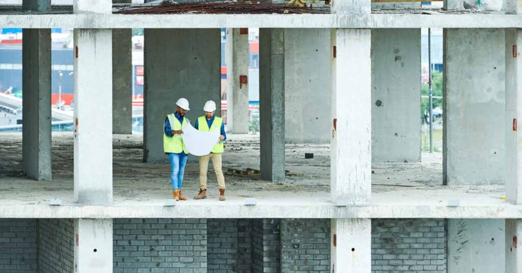 Two people with hardhats and high viz jackets standing in a construction building reading plans