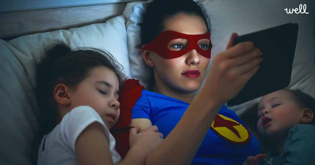 A mother dressed as a superhero lay in bed with 2 infants while she reads a tablet