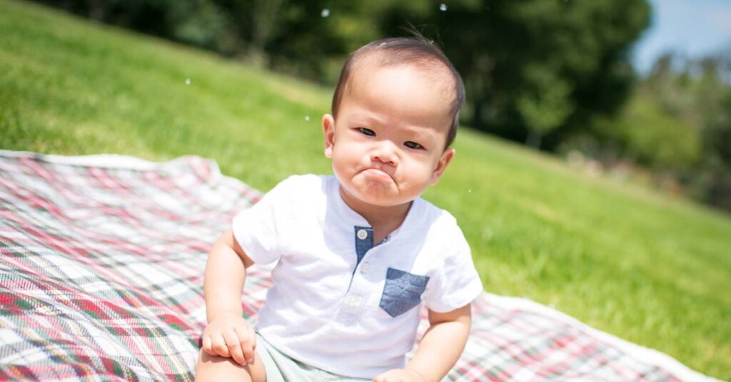 Baby sitting on a picnic rug in the partk with an angry frown