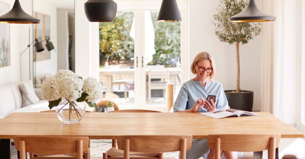 Senior Woman Relaxing With Magazine At Home Looking At Mobile Phone Sitting At Dining Room Table
